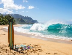 A classic longboard surfboard rests on a sandy beach in front of a peaking Hawaiian wave.