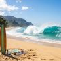 A classic longboard surfboard rests on a sandy beach in front of a peaking Hawaiian wave.
