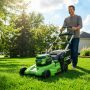 A clean, vibrant shot of a homeowner operating a modern cordless EGO electric lawn mower across a neatly manicured green residential lawn on a bright sunny day.